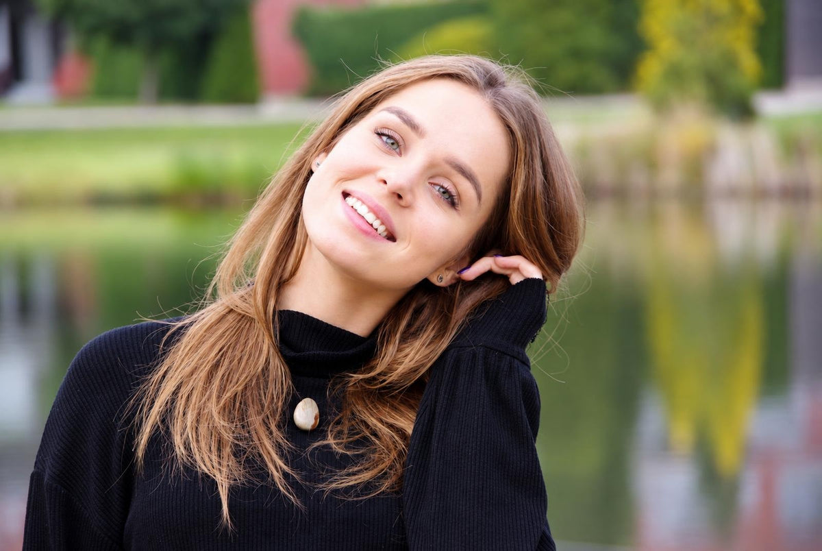 A smiling woman with long brown hair wears a black turtleneck sweater featuring a single light-coloured oval pendant at the neck.