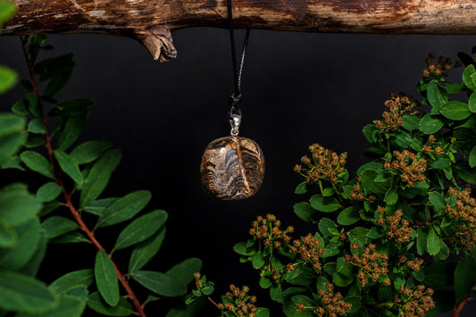 A round, polished pendant with brown and black wavy patterns hangs from a silver bail on a dark cord against a dark background.