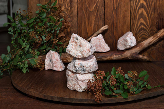 Rough pale pink and gray stones are arranged on a wooden display, with three stacked in front, beside branches and leafy stems.