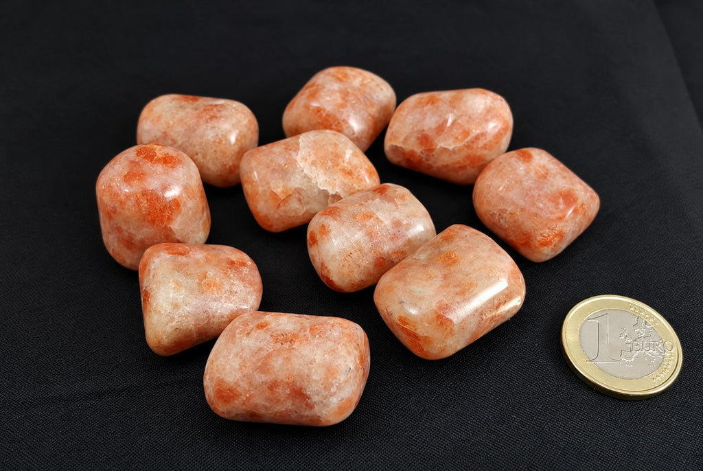 A group of polished, rounded sunstone tumbled stones in orange and cream tones on a dark surface with a one euro coin for scale.