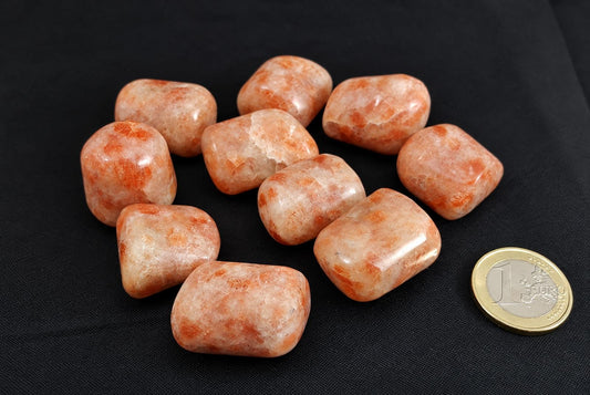 A group of polished, rounded sunstone tumbled stones in orange and cream tones on a dark surface with a one euro coin for scale.