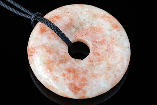 A round, donut-shaped pendant with a central hole, featuring a pale surface with orange and grey speckles, on a dark background.