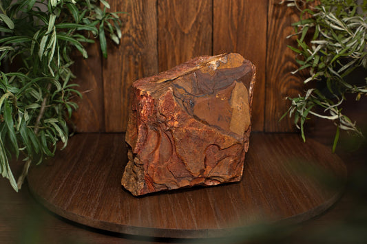A large, rough stone with reddish-brown and golden layers sits on a dark wooden surface, flanked by green plants.