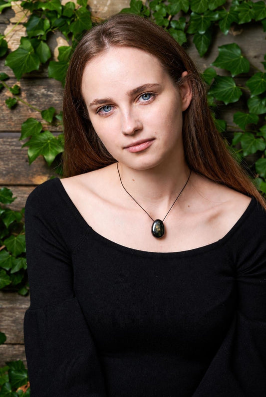 A woman wears a black top and a necklace with a dark, oval-shaped pendant on a thin cord against a wooden background with green ivy.