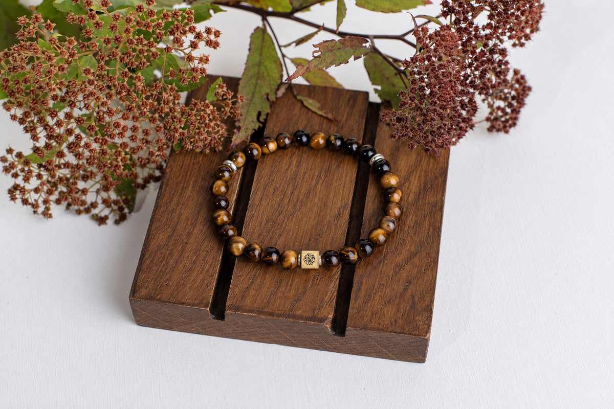 A beaded bracelet with tiger eye and black beads, a square gold-toned charm, and silver spacers rests on a dark wooden block.