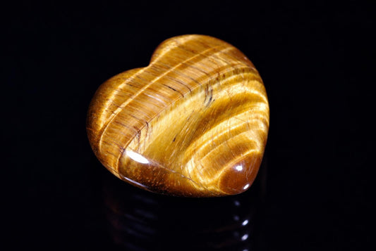 A polished heart-shaped tiger eye stone with golden-brown bands and a glossy finish on a dark surface.