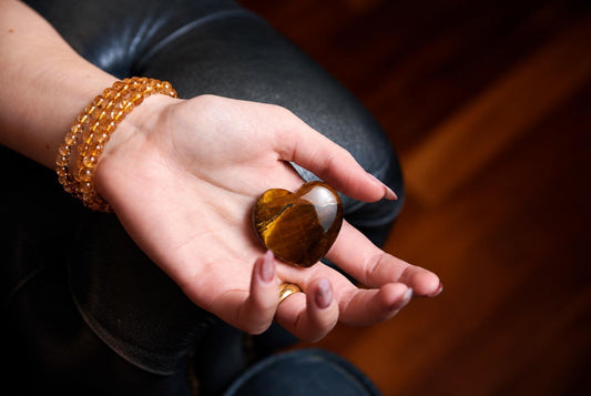 A hand holds a polished, heart-shaped tiger eye stone with golden-brown stripes on a dark surface.