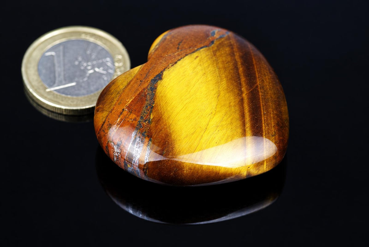 A polished heart-shaped stone with yellow and brown stripes sits on a dark surface next to a coin.