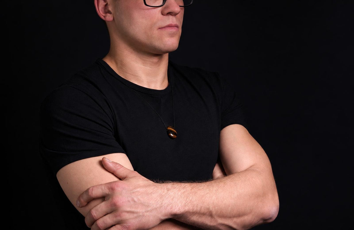 A man in a black shirt wears a small brown oval pendant on a dark cord, with his arms crossed against a dark background.