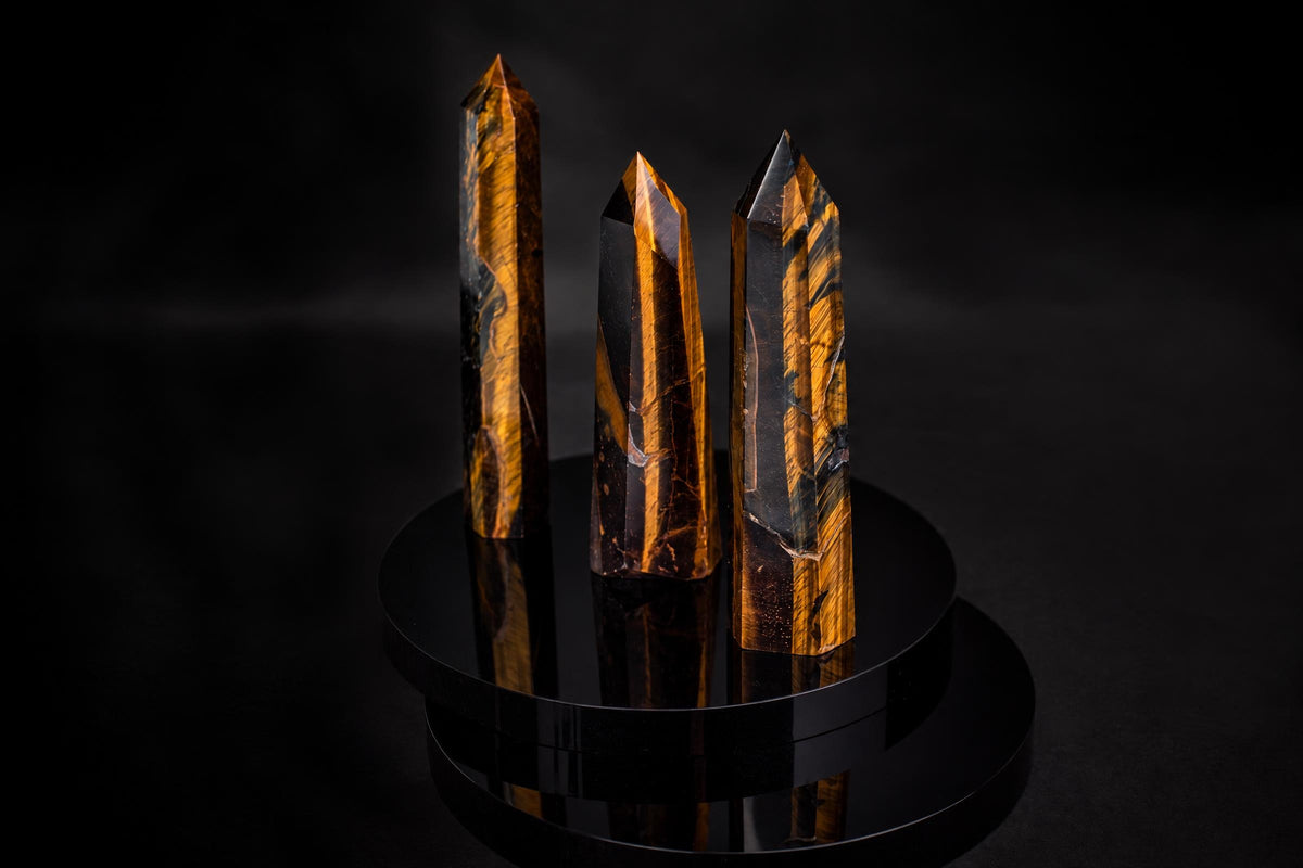 Three polished tiger eye obelisks with golden-brown stripes stand on a dark, reflective circular base against a black background.
