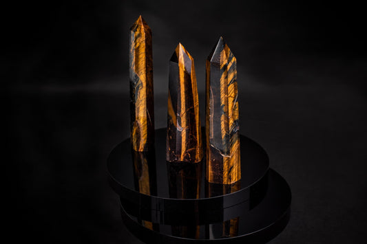 Three polished tiger eye obelisks with golden-brown stripes stand on a dark, reflective circular base against a black background.