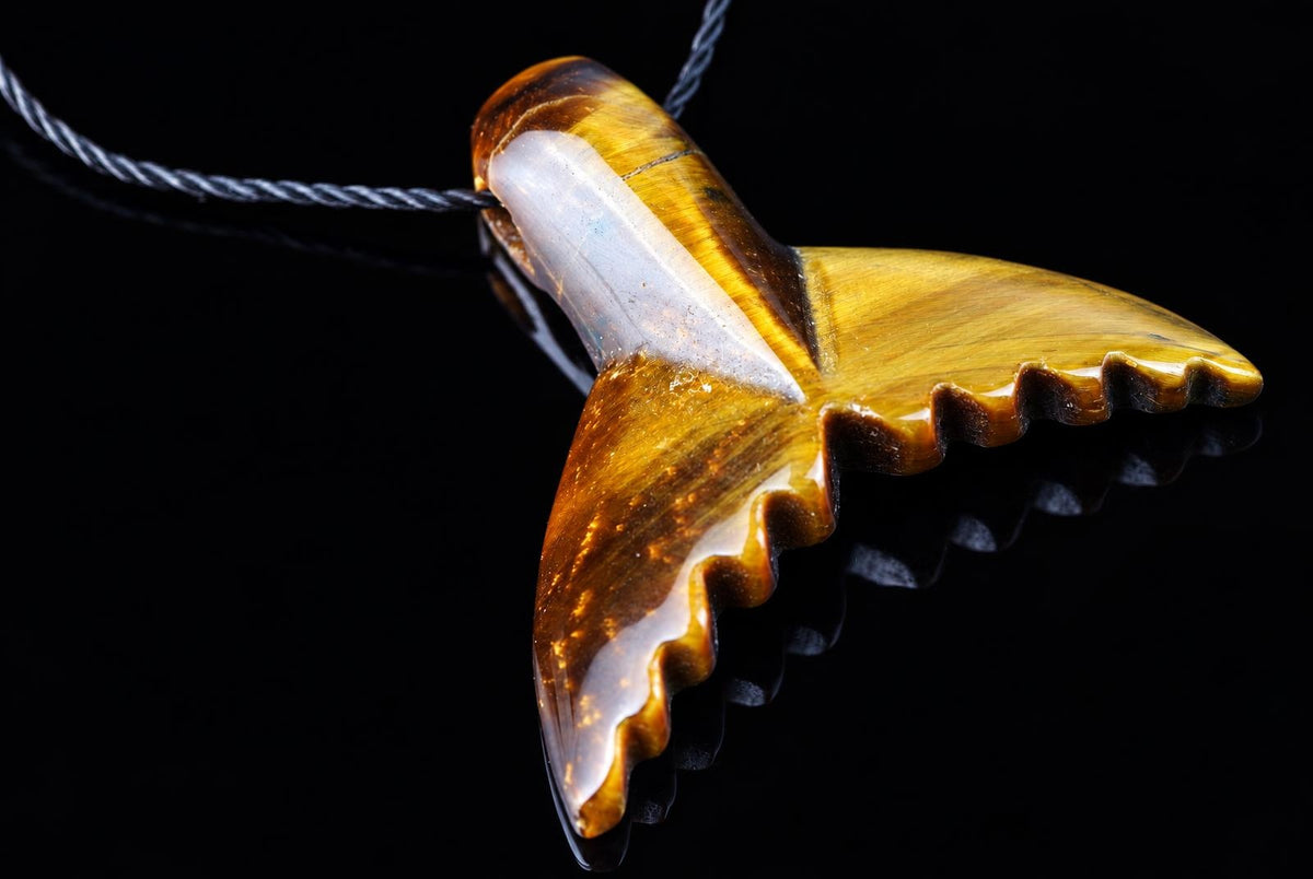 A polished pendant shaped like a dolphin tail, featuring golden-brown tiger eye stone with white inclusions, hanging from a dark cord against a black background.