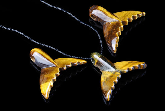 Three polished yellow-brown pendants shaped like dolphin tails with scalloped edges, each on a grey cord against a dark reflective surface.