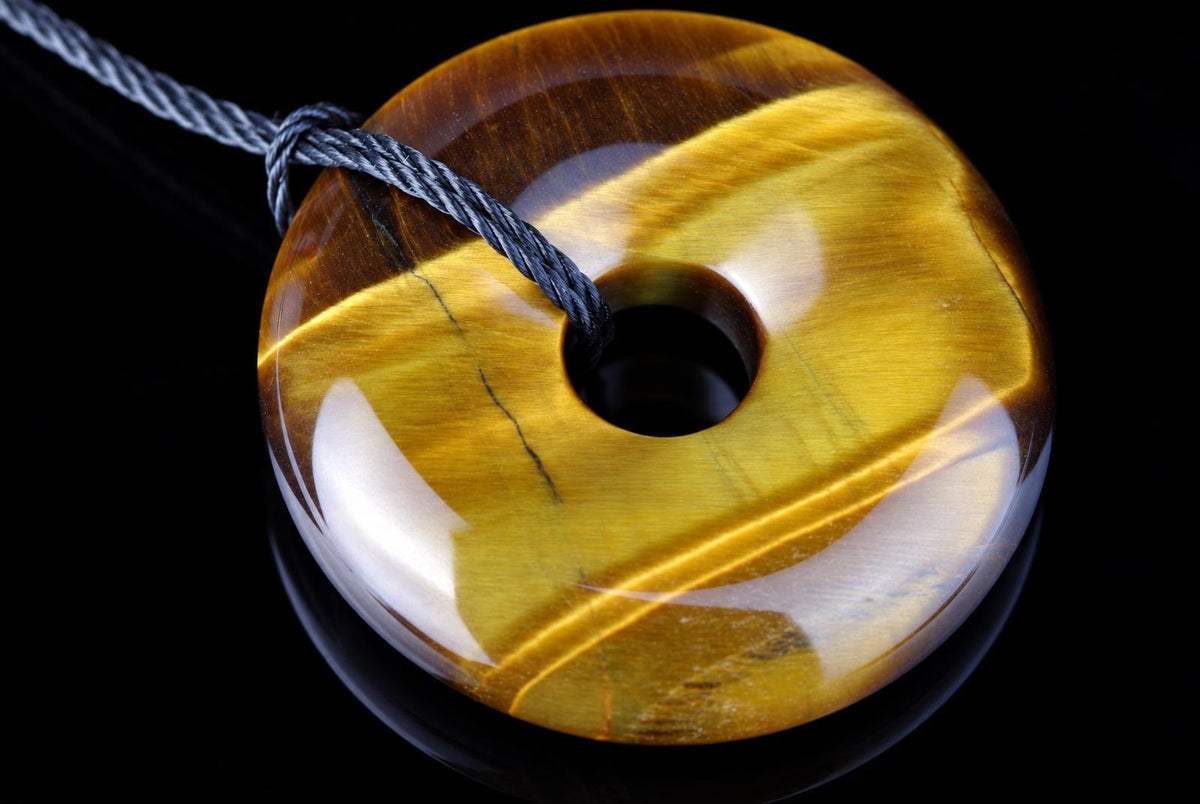 A polished, donut-shaped pendant with golden-brown tiger eye stripes and a central hole, threaded on a grey cord against a dark surface.