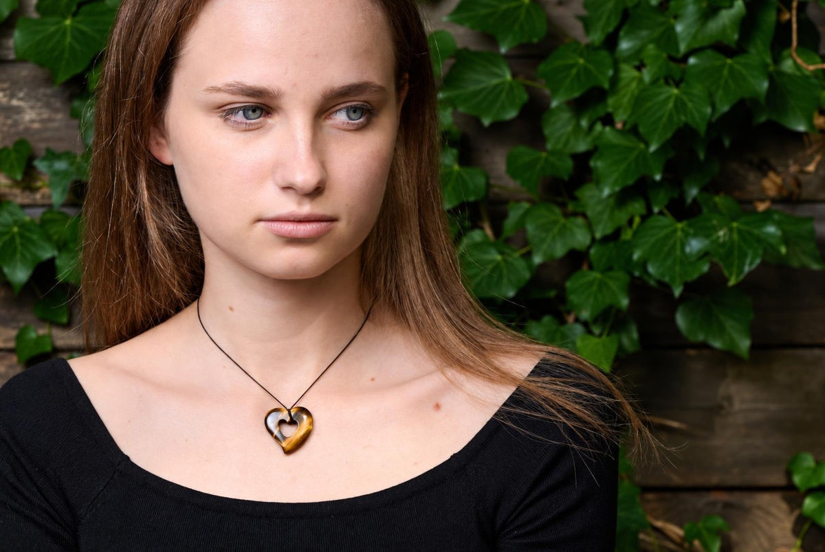 A woman wears a heart-shaped pendant with tiger eye stripes on a black cord, against green ivy and wood.