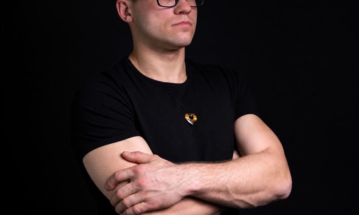 A man in a black shirt wears a small brown heart-shaped pendant on a dark cord, standing with his arms crossed.