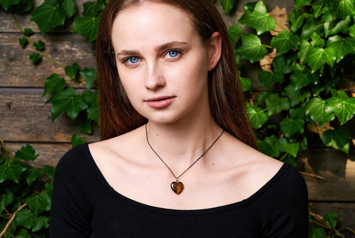 A woman wears a black top and a necklace with a heart-shaped brown pendant on a dark cord, against a wooden wall with green ivy.