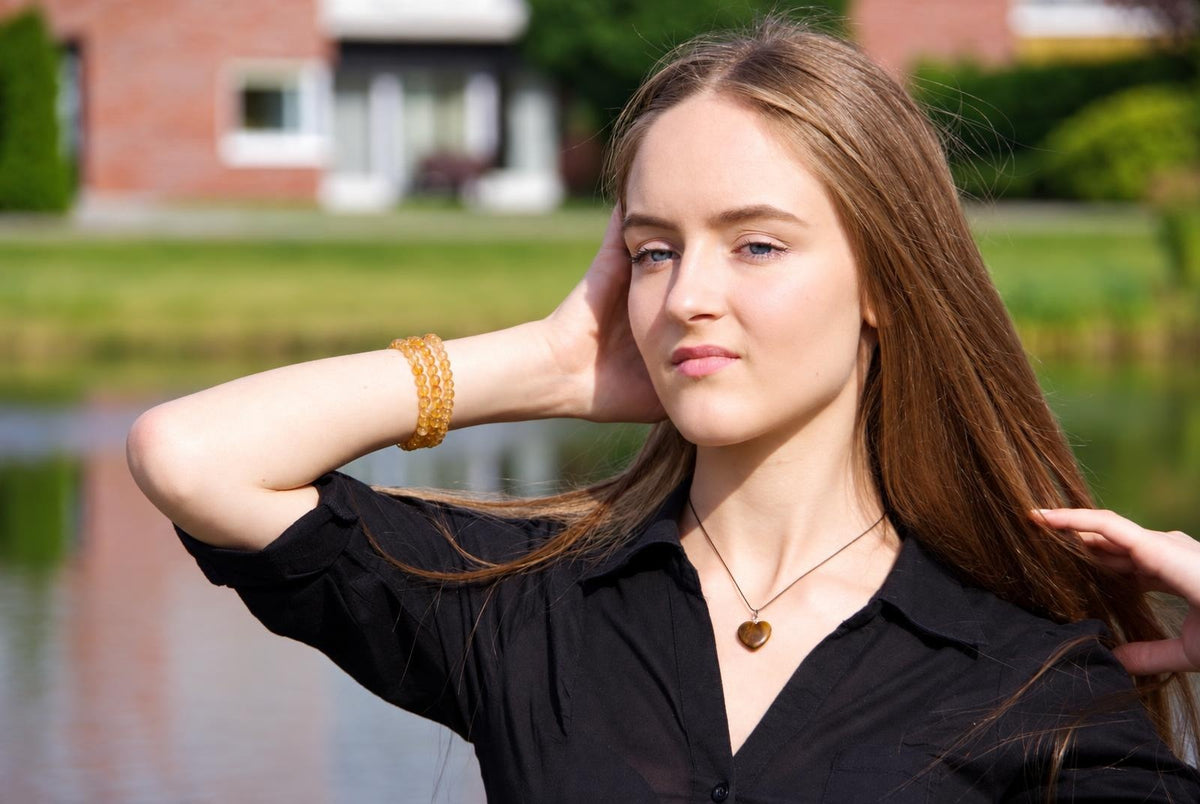 A woman in a black shirt wears a small brown heart pendant outdoors, with one arm raised and blurred greenery behind her.