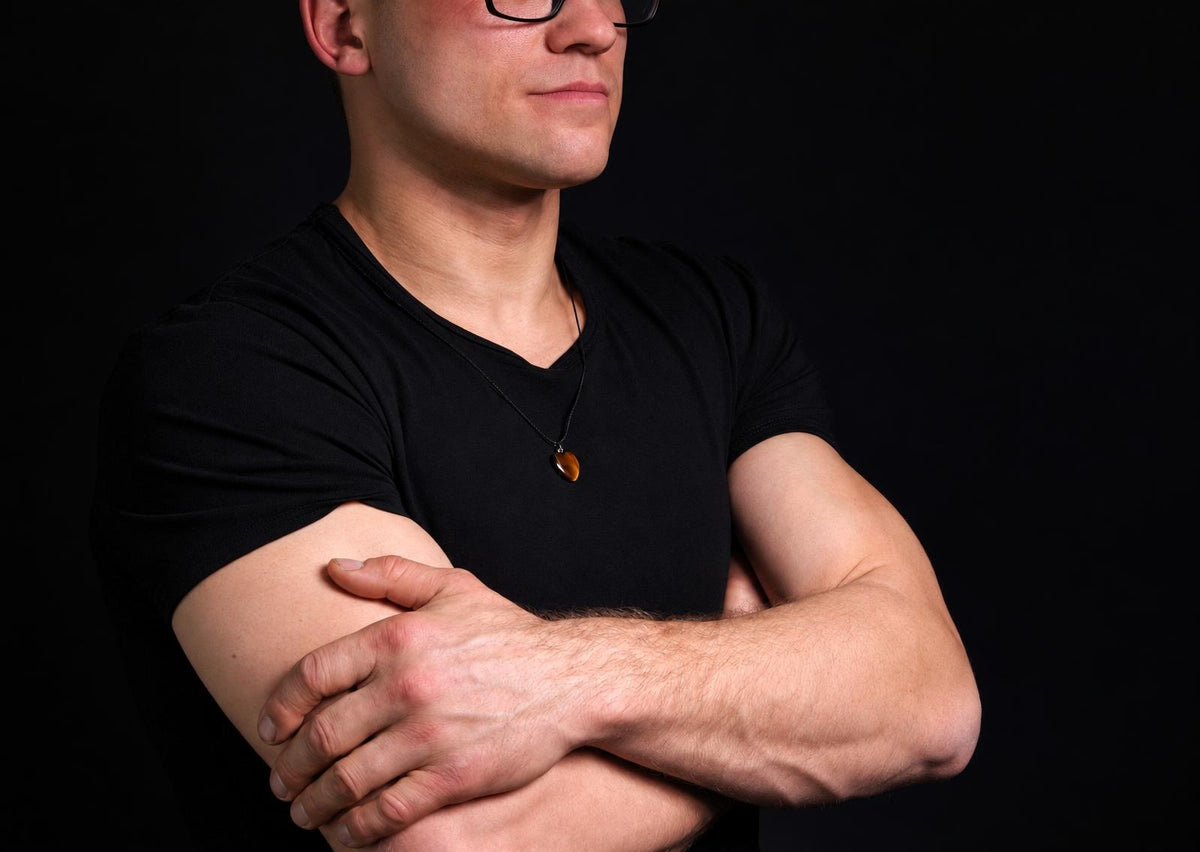 A man with folded arms wears a small brown tiger eye heart pendant on a black cord against his black shirt.