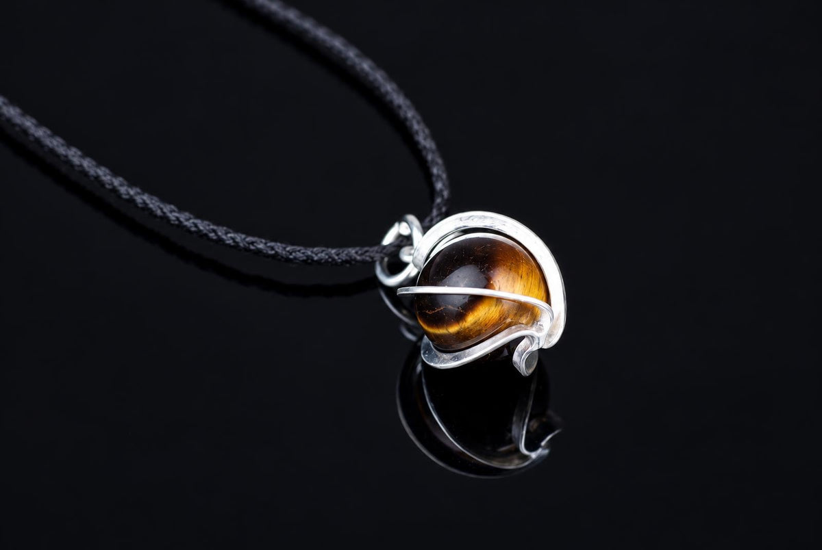 A small brown stone sphere with gold-colored bands sits inside a curved metal setting on a black cord against a reflective black surface.