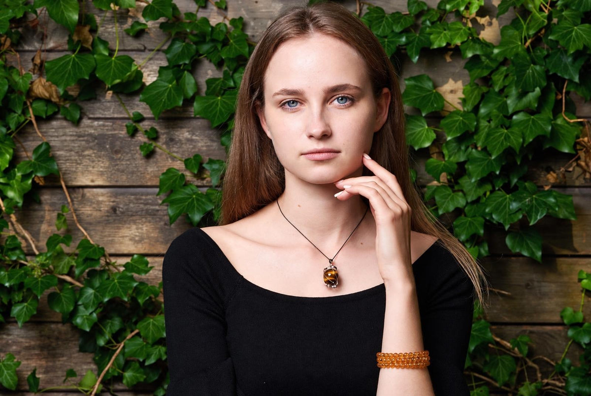 A woman wears a rounded brown tiger eye pendant on a black cord, standing before a wooden wall covered with green leaves.