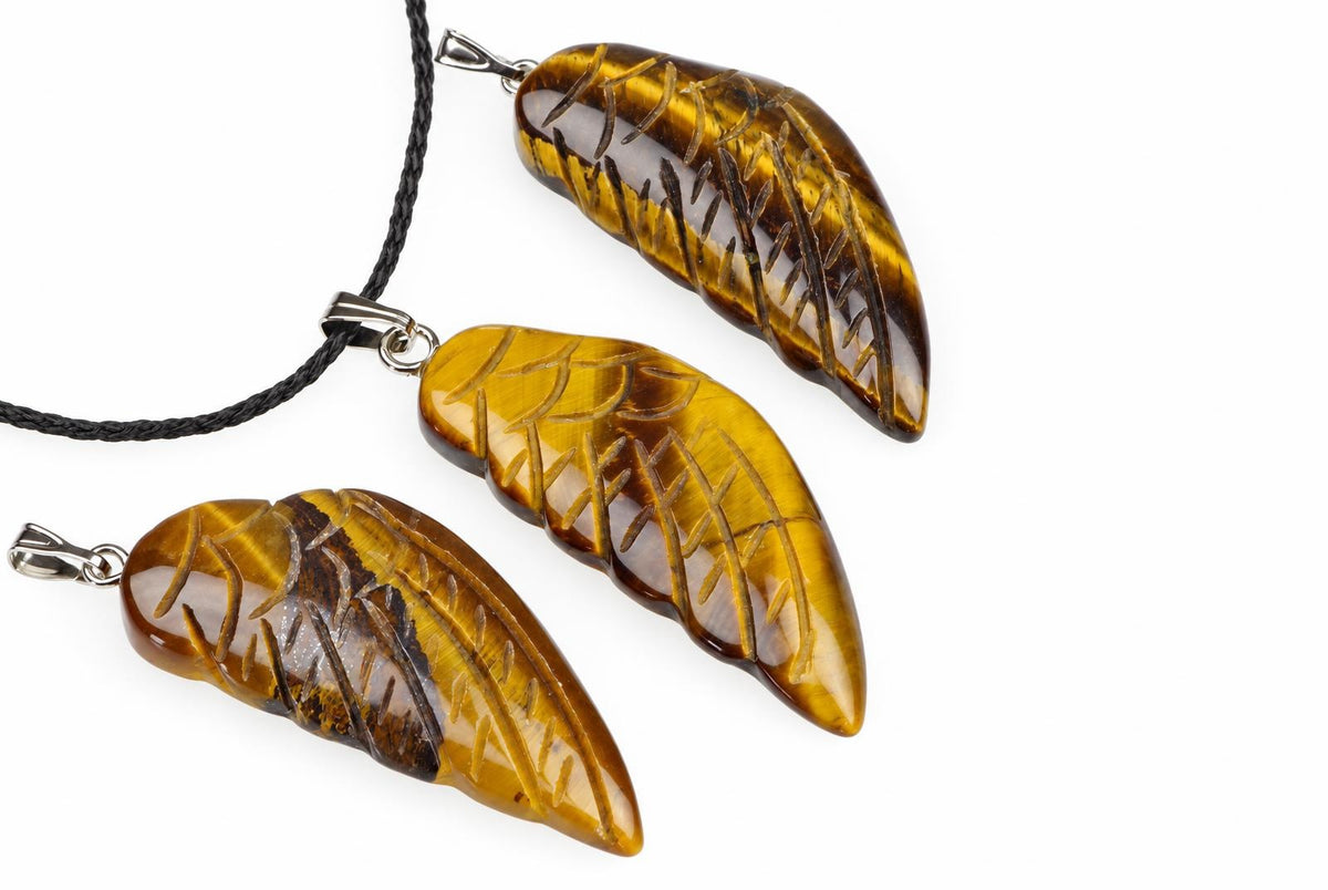 Three carved tiger eye pendants shaped like wings, each with a silver-toned bail and one on a black cord, arranged diagonally on a white surface.