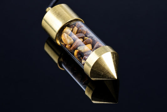A brass pendulum with a clear tube containing brown and yellow stones, resting on a dark reflective surface.