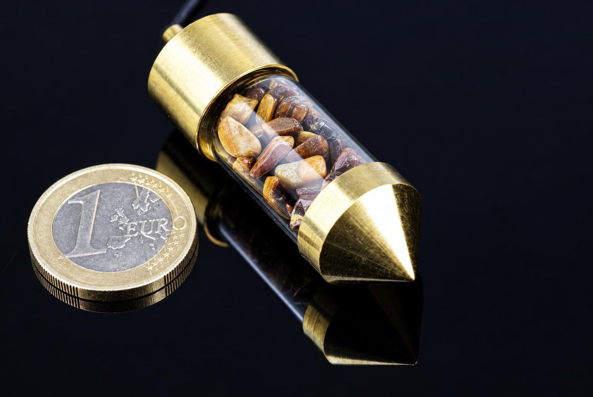 A brass pendulum with a clear tube filled with brown and yellow stones, next to a one-euro coin on a dark surface.