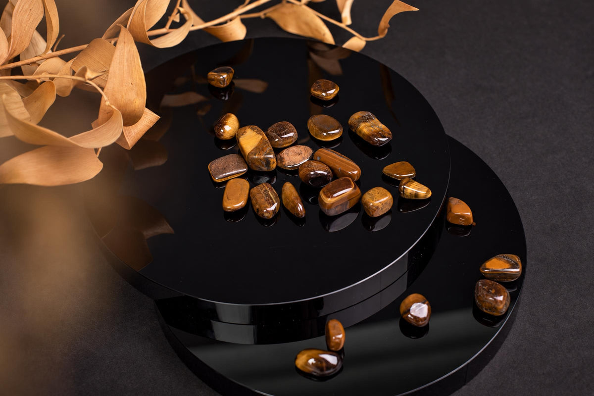 Small polished brown stones with gold-colored bands are scattered over a black circular display and nearby surface beneath dried leaves.