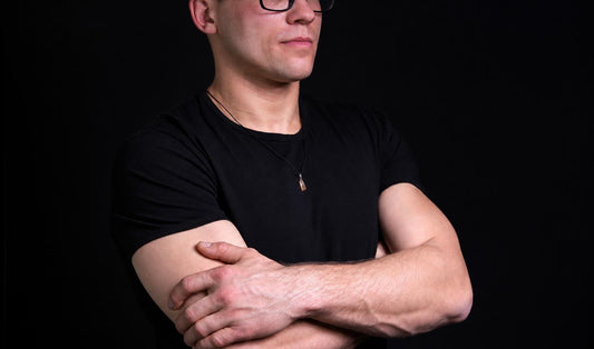 A man in a black t-shirt wears a necklace with a small, golden-yellow pendant against a dark background.