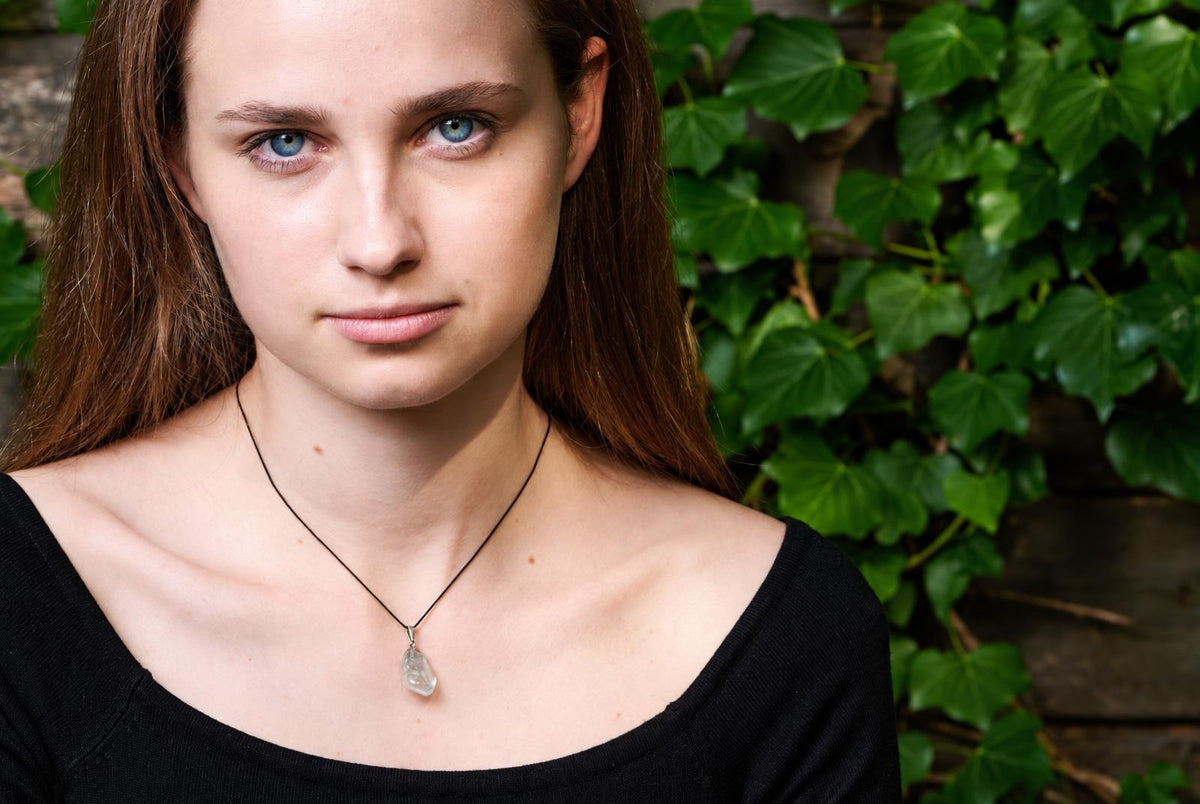 A woman wears a necklace with a clear, teardrop-shaped pendant on a dark cord against green foliage.