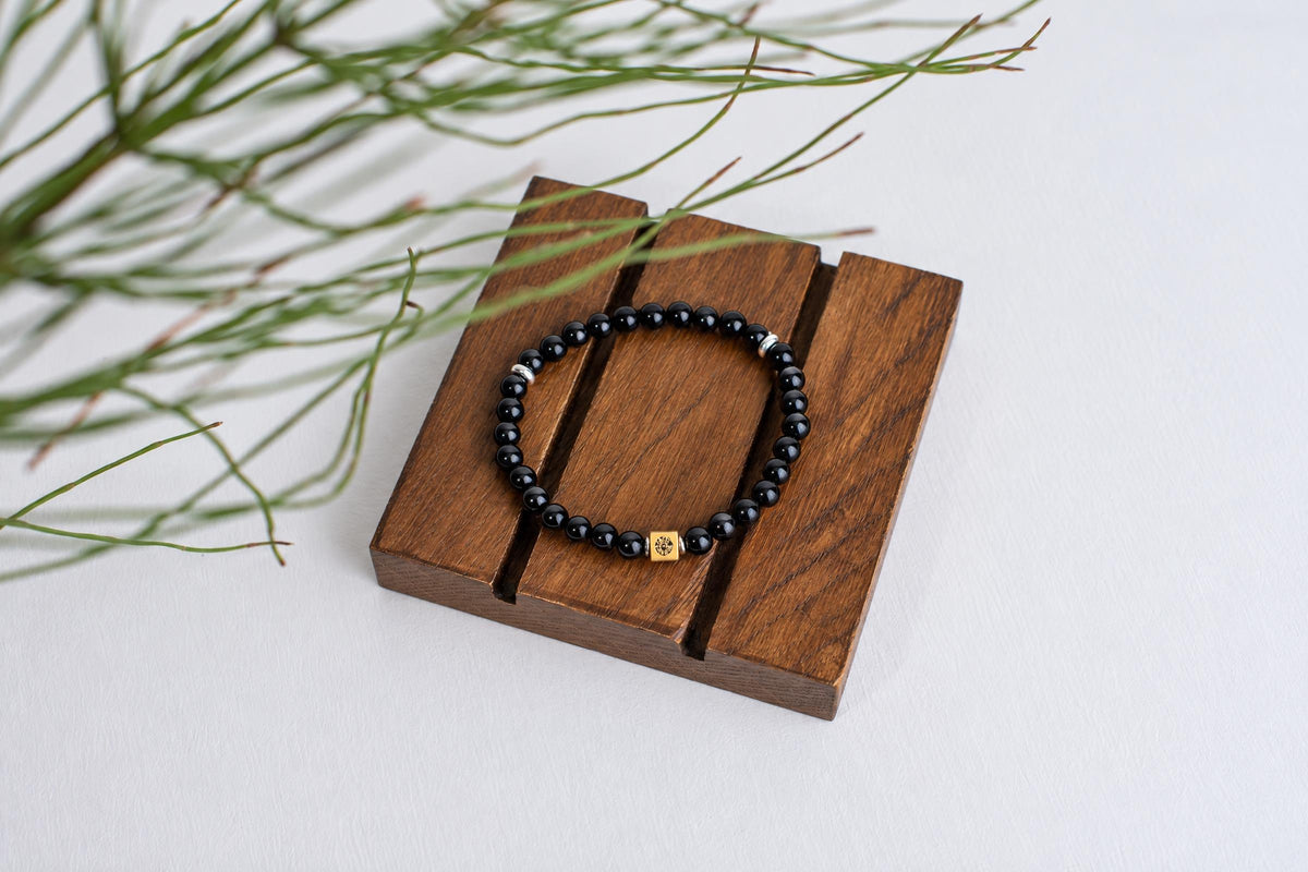 A black beaded bracelet with a gold-toned square charm rests on a dark wooden surface, next to green foliage.