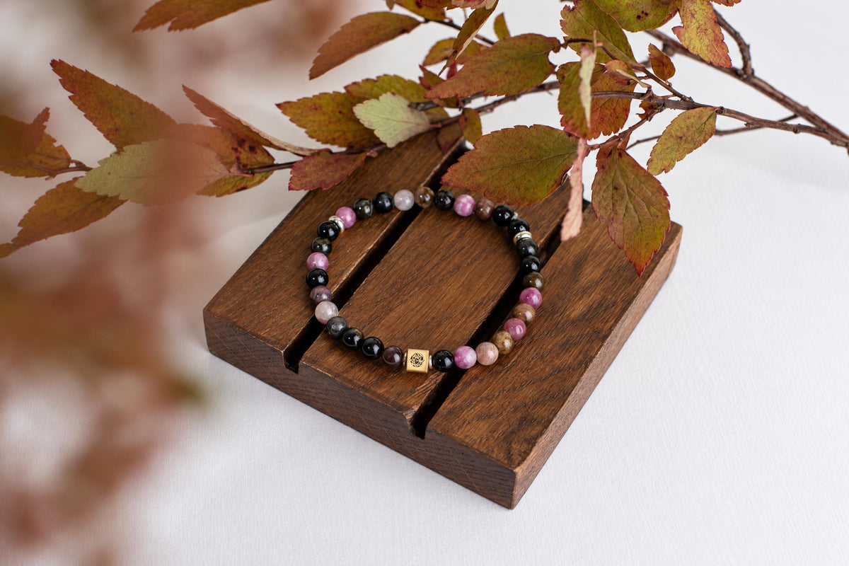 A beaded bracelet with black, pink, and brown beads sits on a dark wooden surface, partially framed by autumn leaves.