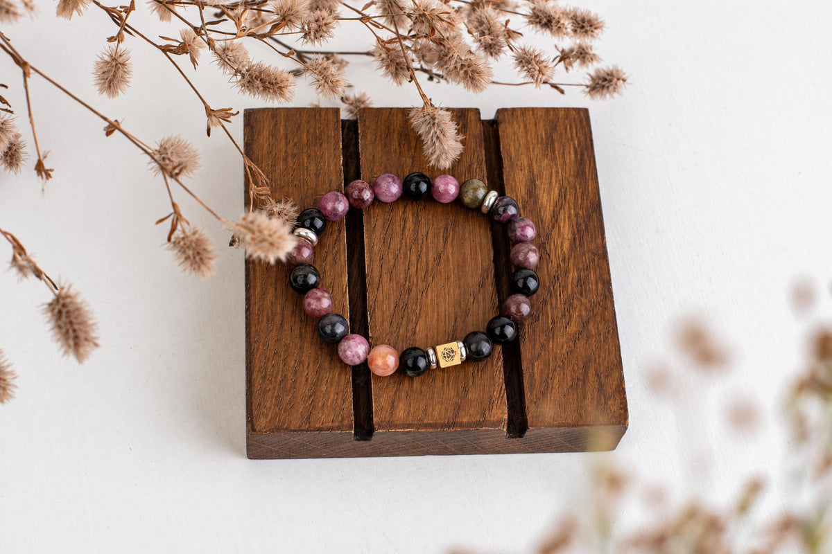 A beaded bracelet with pink, black, and green round beads on a dark wooden stand, surrounded by dried flowers.