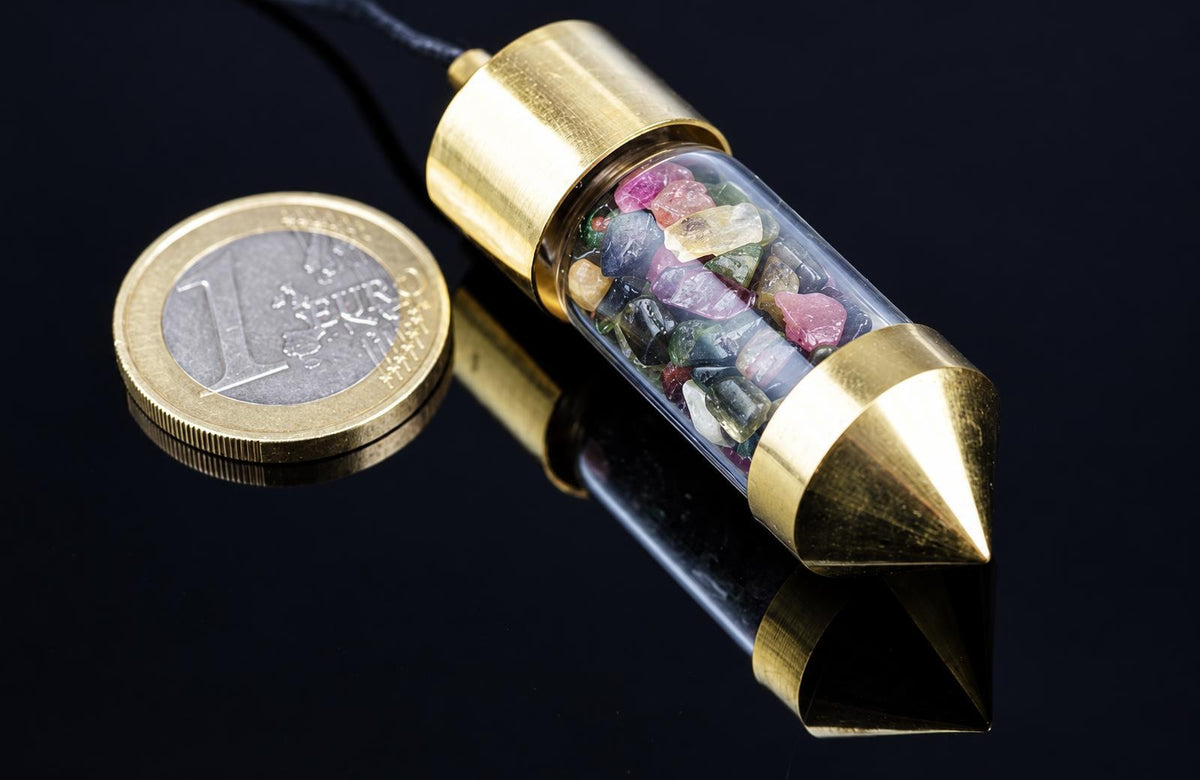 A brass pendulum with a clear tube containing multi-coloured tourmaline stones, next to a one-euro coin on a dark surface.