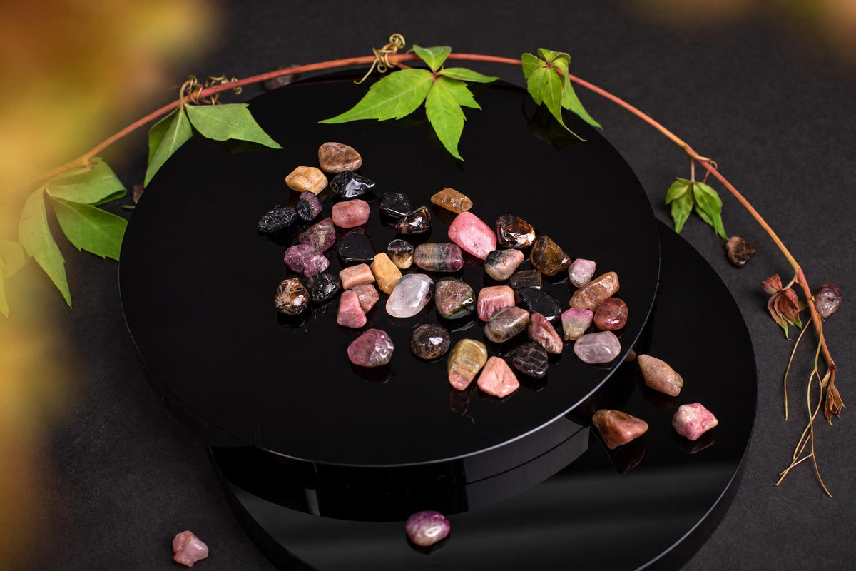 A collection of multi-colored tourmaline tumbled stones arranged on a glossy black circular surface, surrounded by green leaves and a vine.