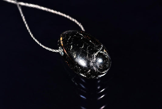 A polished black drop-shaped pendant with pale fine markings rests with its cord on a reflective black surface.