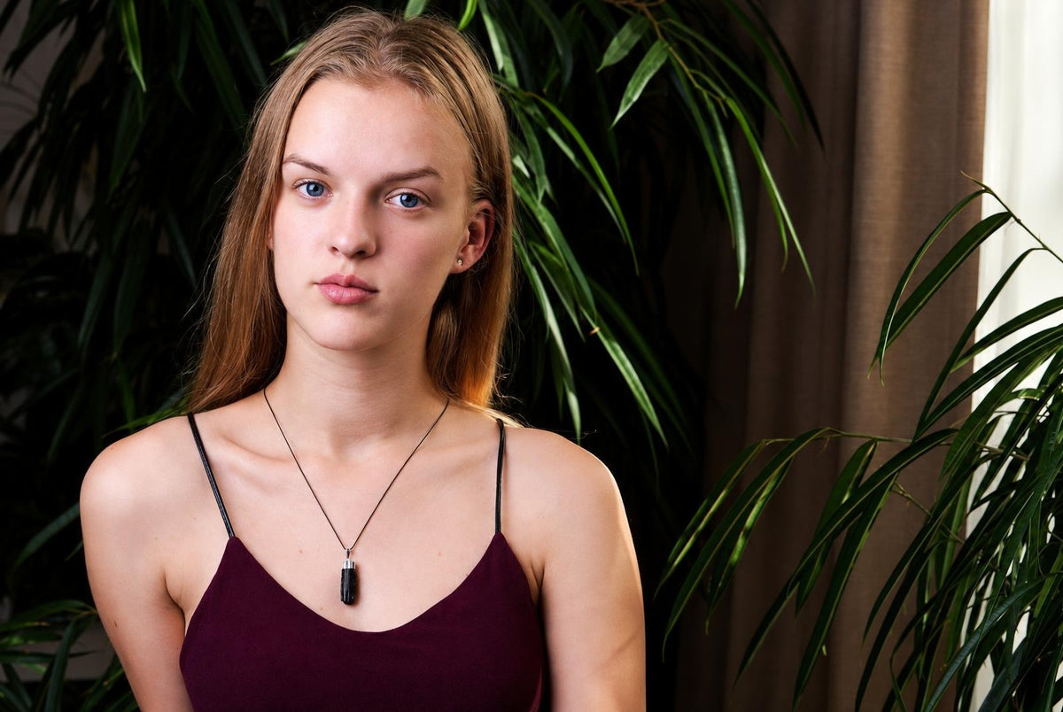 A woman wears a dark maroon top and a necklace with a black, cylindrical pendant on a thin cord.