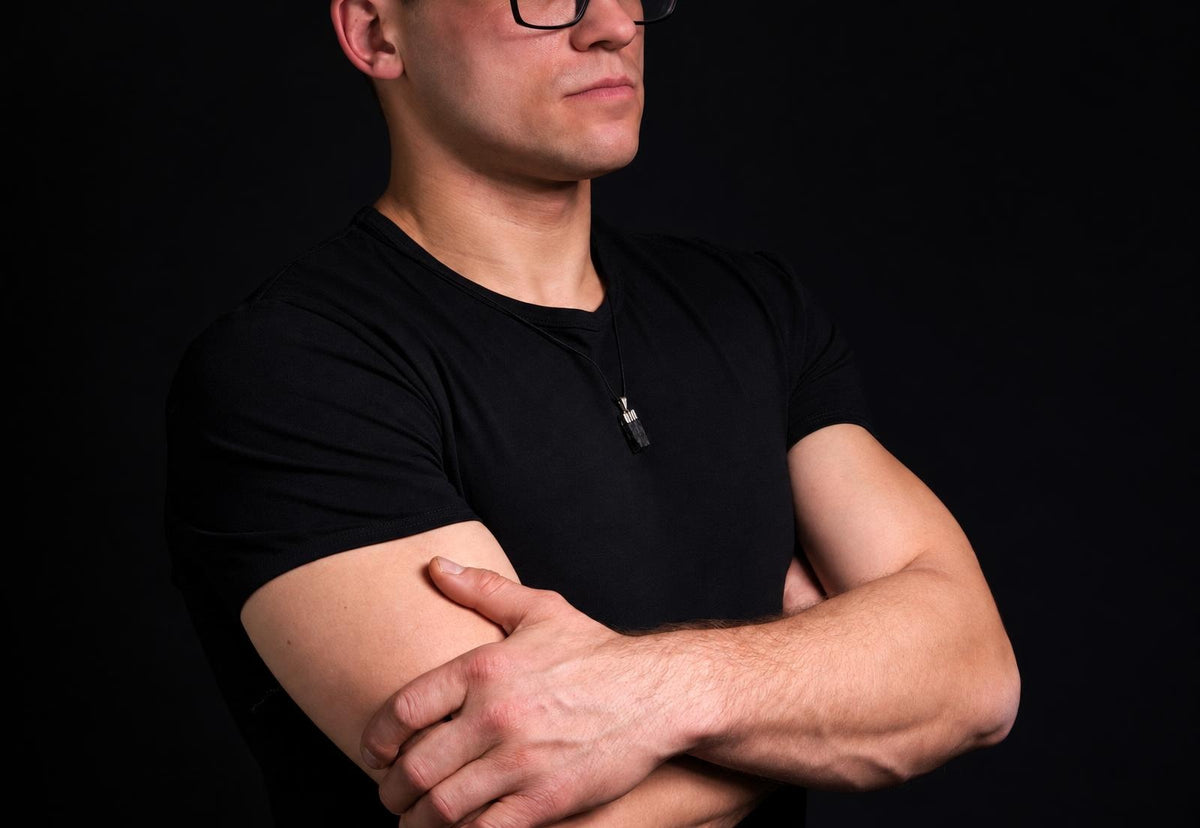 A man in a black t-shirt wears a small dark pendant on a thin cord against a dark background.
