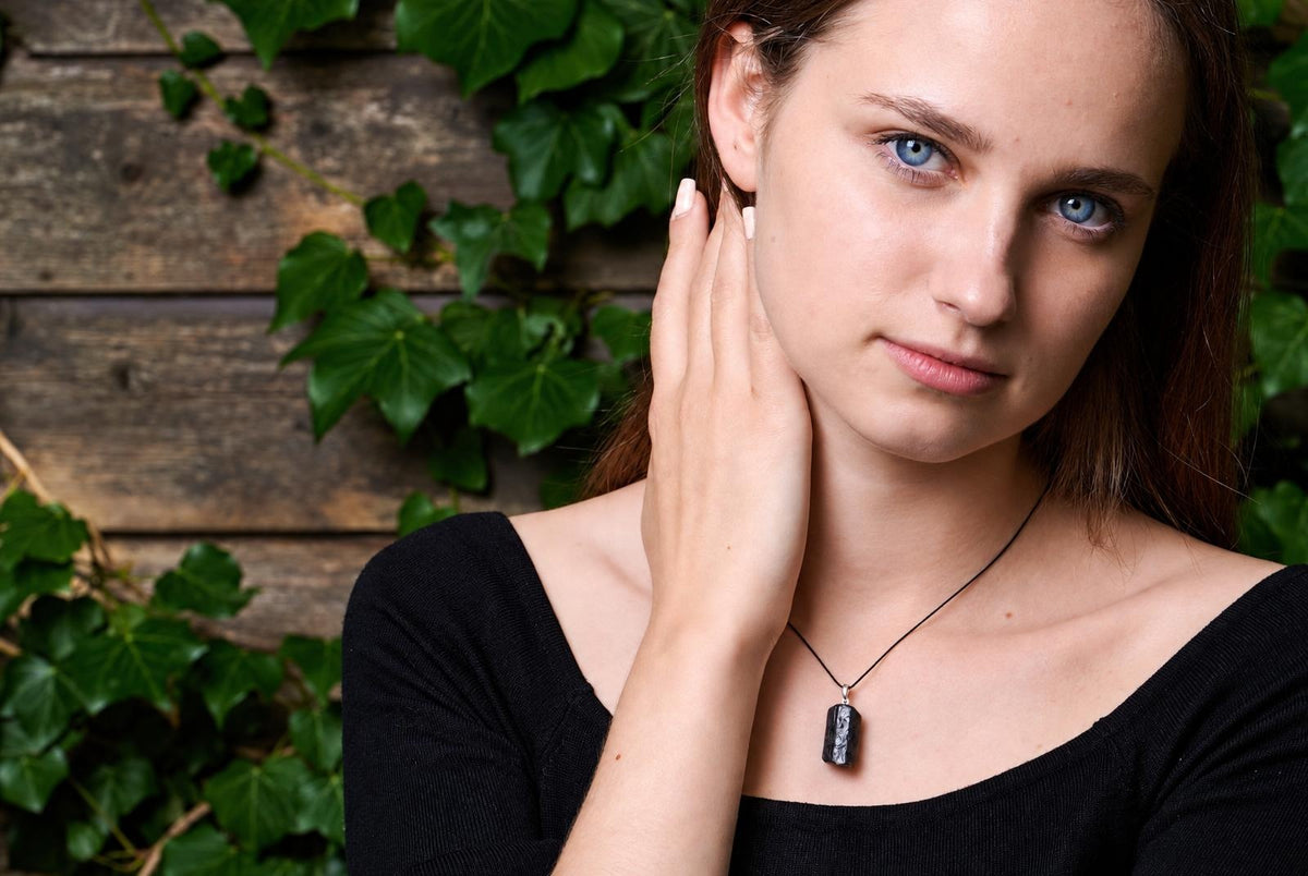 A woman wears a black top and a dark, rectangular pendant on a thin cord against a wooden wall with green ivy.