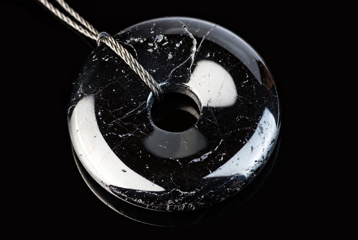 A polished black donut-shaped pendant with white inclusions and a central hole, suspended on a silver-toned cord against a dark surface.