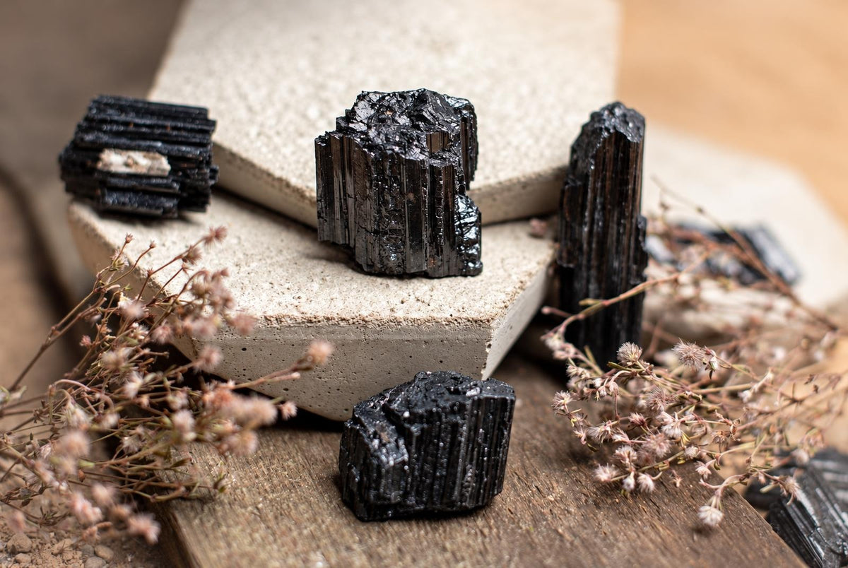 Several dark, metallic black Tourmaline schorl crystals with vertical striations rest on light concrete blocks and a wooden surface, surrounded by dried flowers.