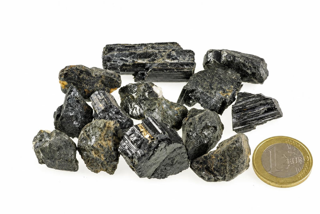 A collection of dark, black tourmaline schorl crystals with some gold-coloured inclusions, arranged on a white surface next to a one-euro coin.