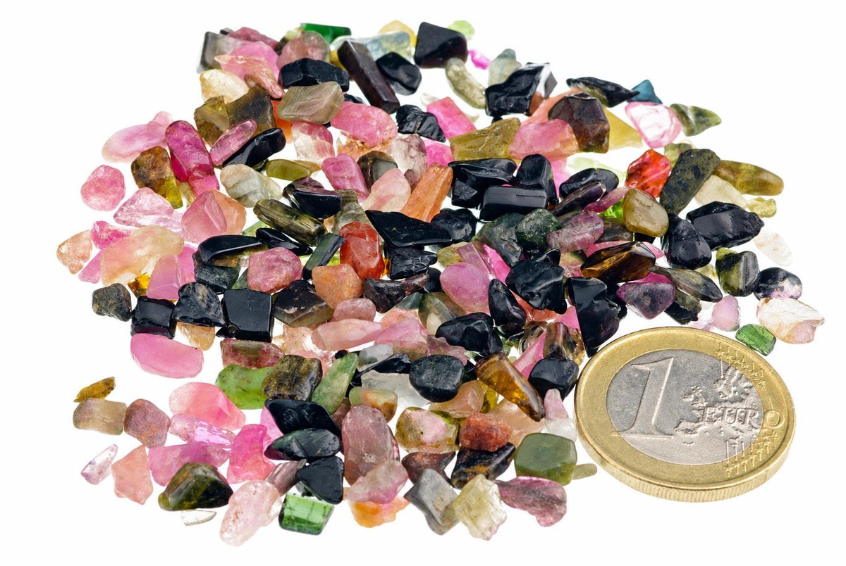 A pile of small, rough tourmaline stones in pink, black, green and orange next to a one euro coin on white.