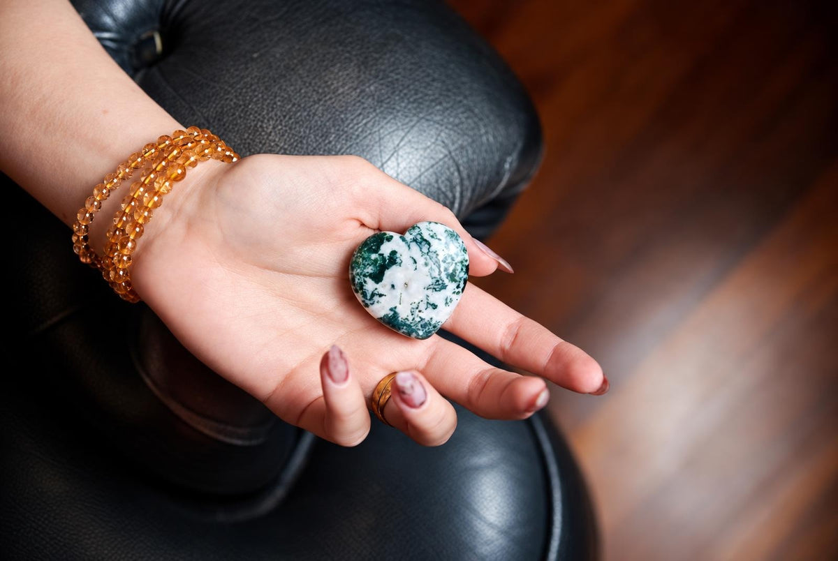A hand holds a polished heart-shaped stone with green and white patterns, resting on a dark leather surface.