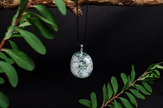A polished, oval-shaped pendant with white and green speckled stone hangs from a black cord on a dark surface.