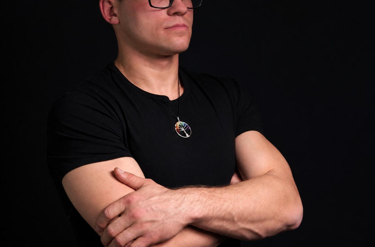 A man wearing a black shirt and glasses displays a small circular tree of life pendant, standing with his arms crossed.