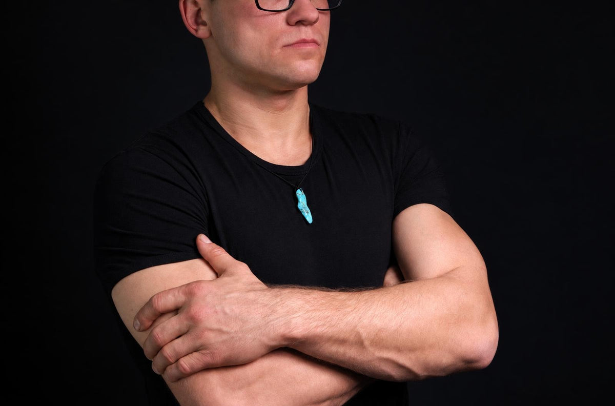 A man in a black t-shirt wears a turquoise pendant on a dark surface against a black background.