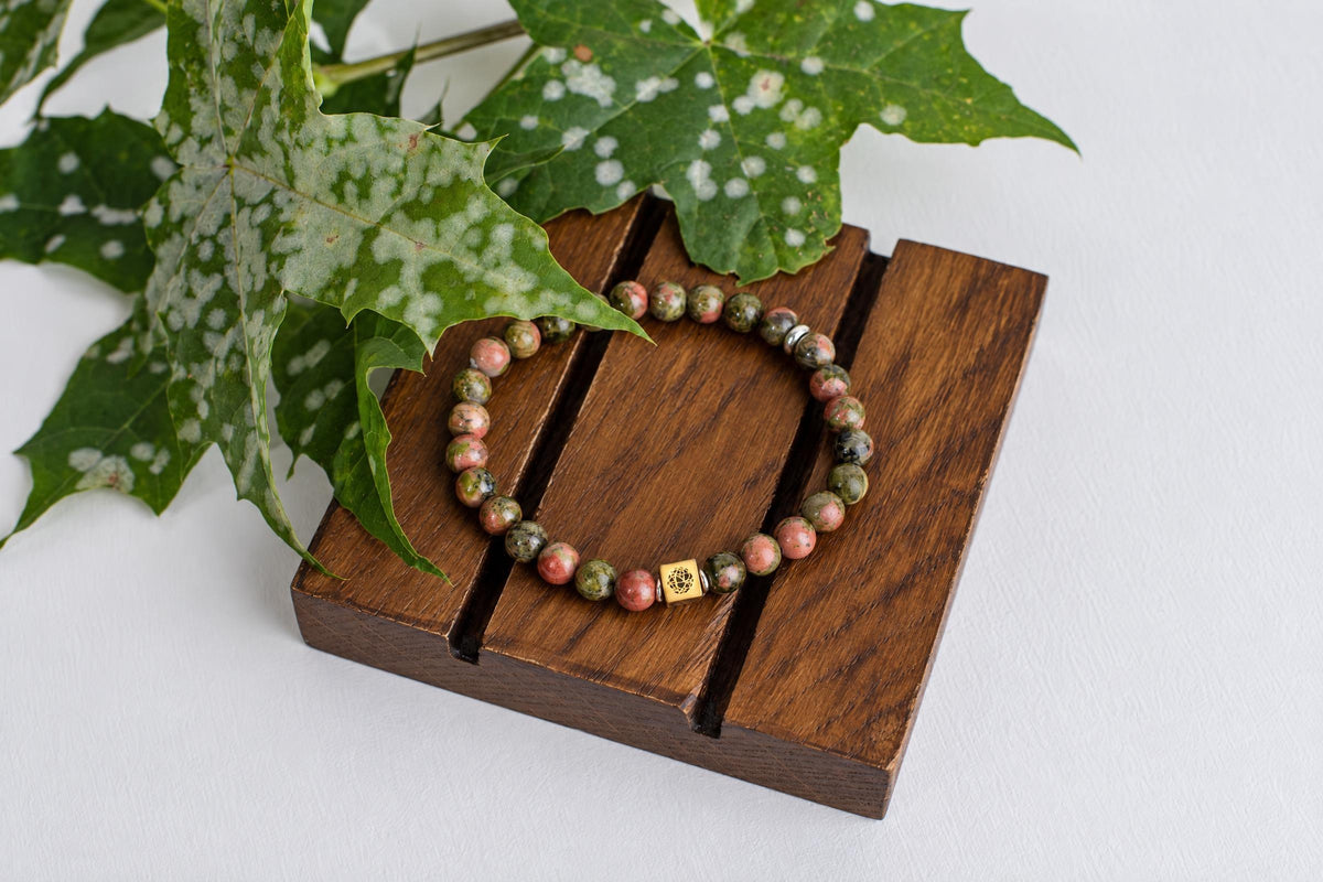 A beaded bracelet with pink and green stones rests on a dark wooden surface, accompanied by green leaves.
