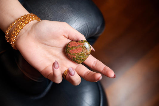 A hand holds a polished heart-shaped stone with red and green patterns on a dark surface.